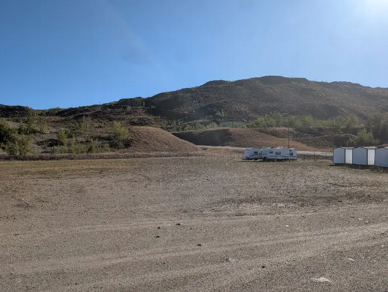 The open gravel yard with a travel trailer parked along the fence line and sheds to the right